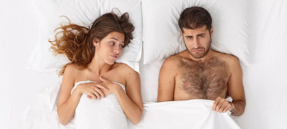 A couple are lying in bed, frowning